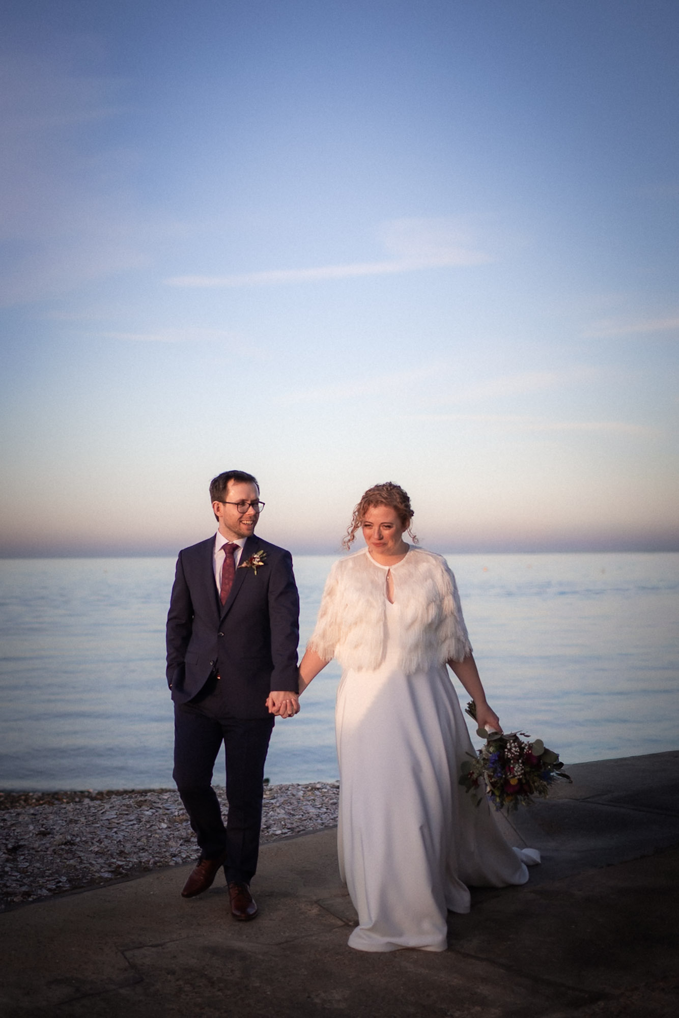 Bride and groom walking hand in hand along a waterfront at sunset, with soft pastel light reflecting off the water.