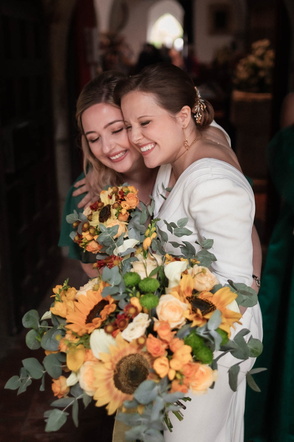 Bride and bridesmaid embracing at the entrance of a church, holding vibrant sunflower and rose bouquets during a joyful wedding moment.”