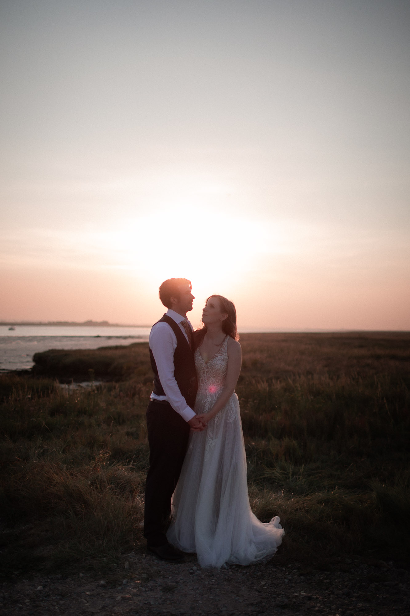 Bride and groom standing hand in hand at sunset near the water, softly backlit by the golden sky.