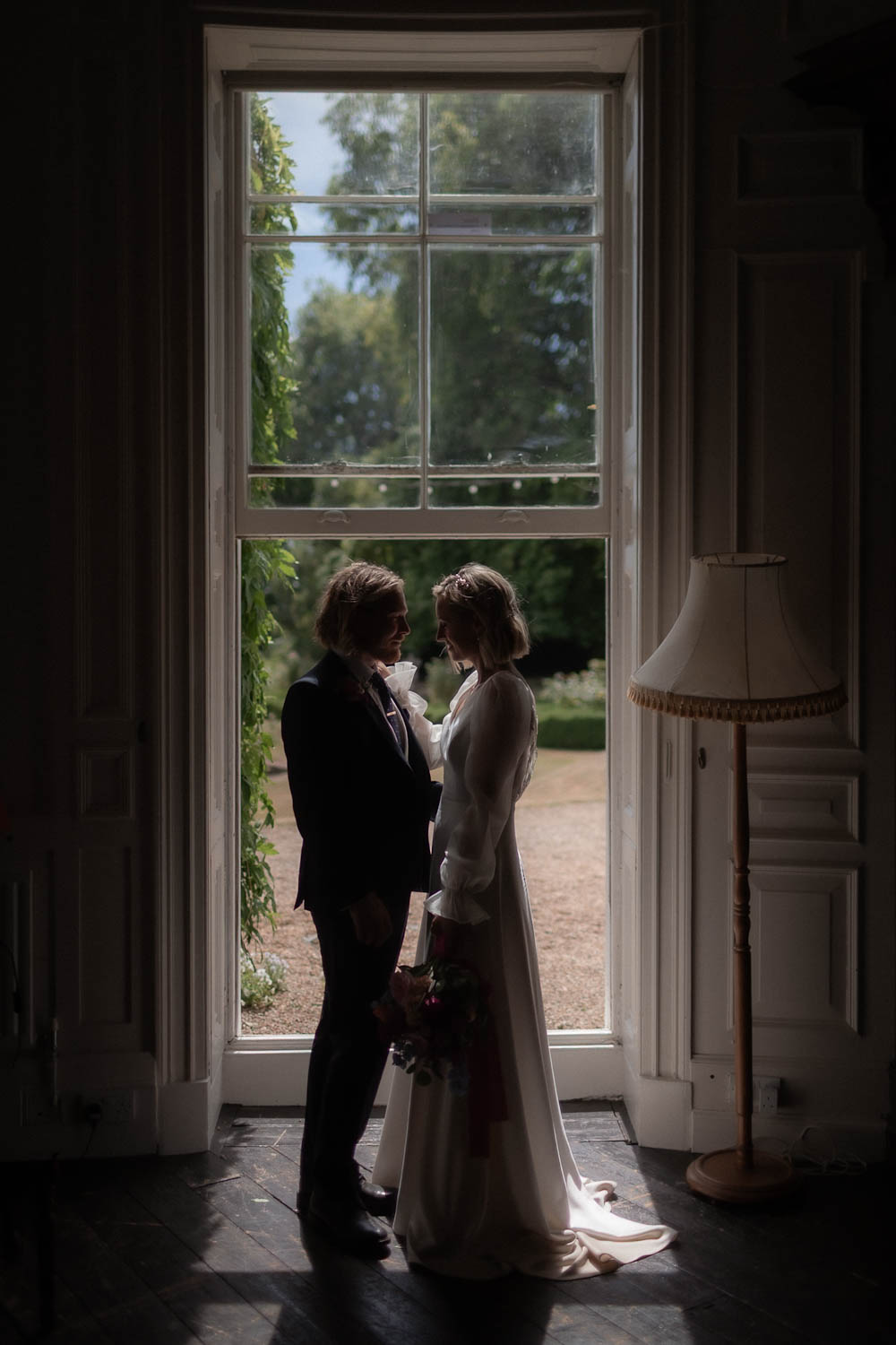 Silhouetted bride and groom standing together by a large window, creating a warm, intimate wedding moment