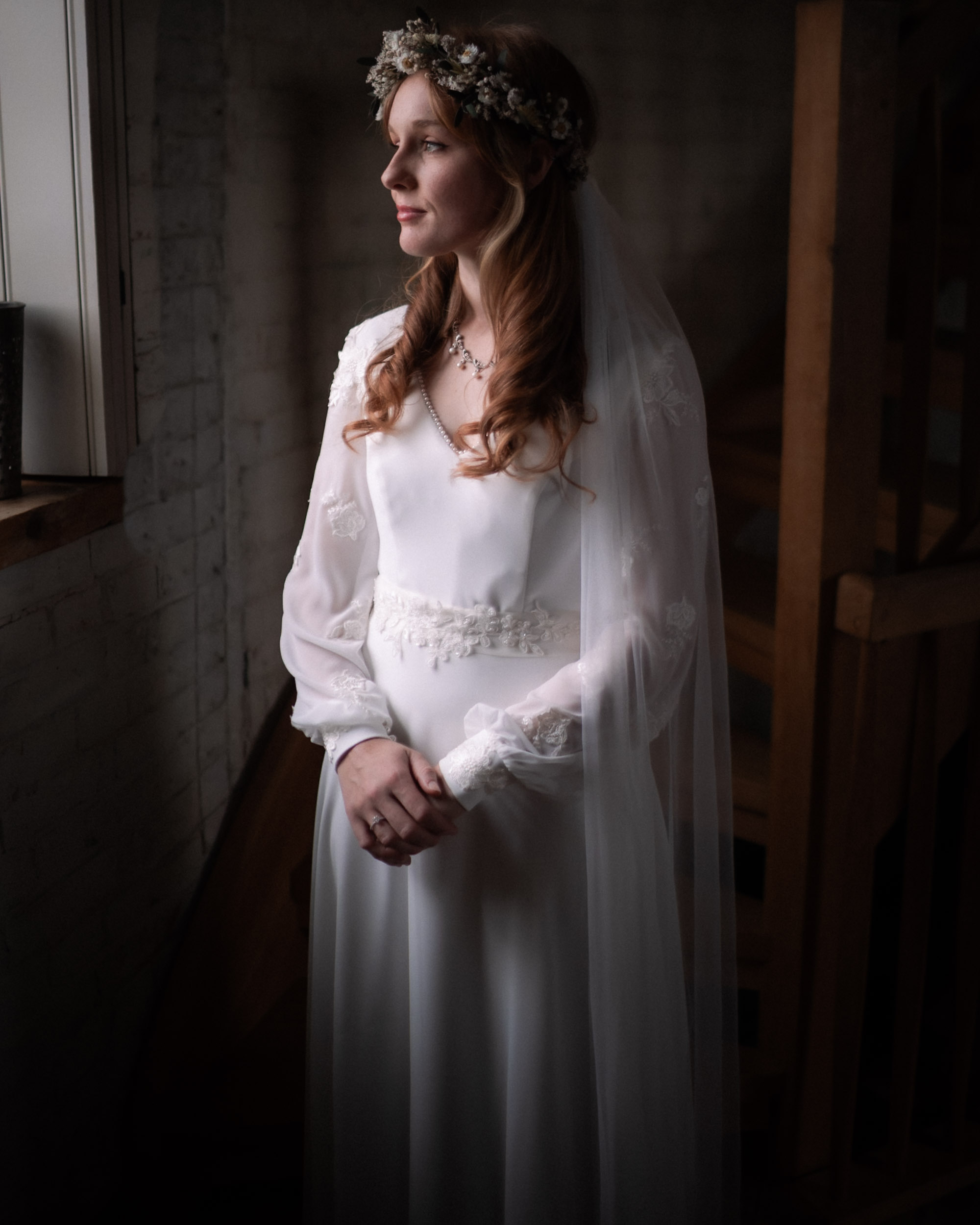 Bride standing by a window in a lace gown and floral crown, softly lit at the entrance of a church.