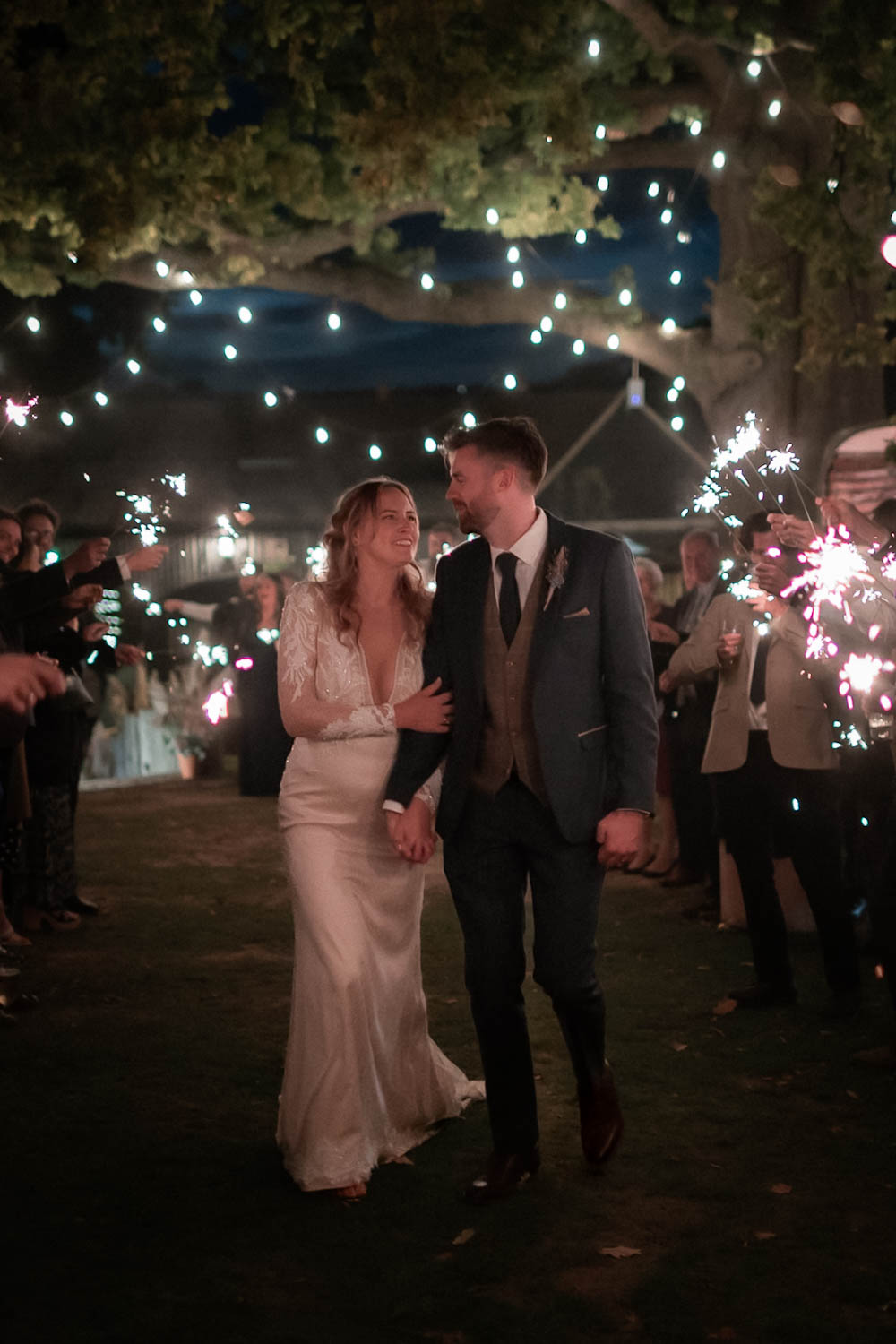 Bride and groom walking through a sparkler tunnel at night under hanging lights, surrounded by cheering guests.