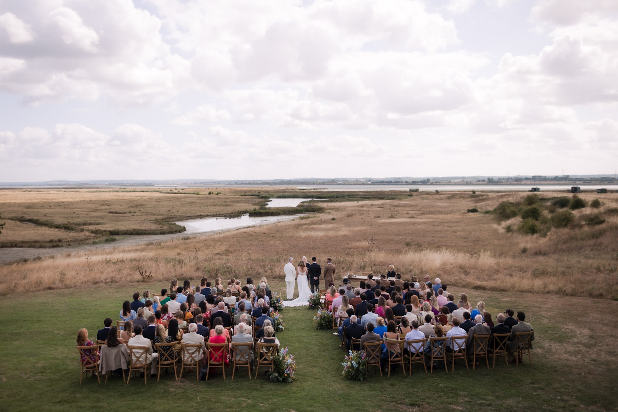 Outdoor wedding ceremony in a wide open field, with guests seated and a couple standing at the altar beside a river.