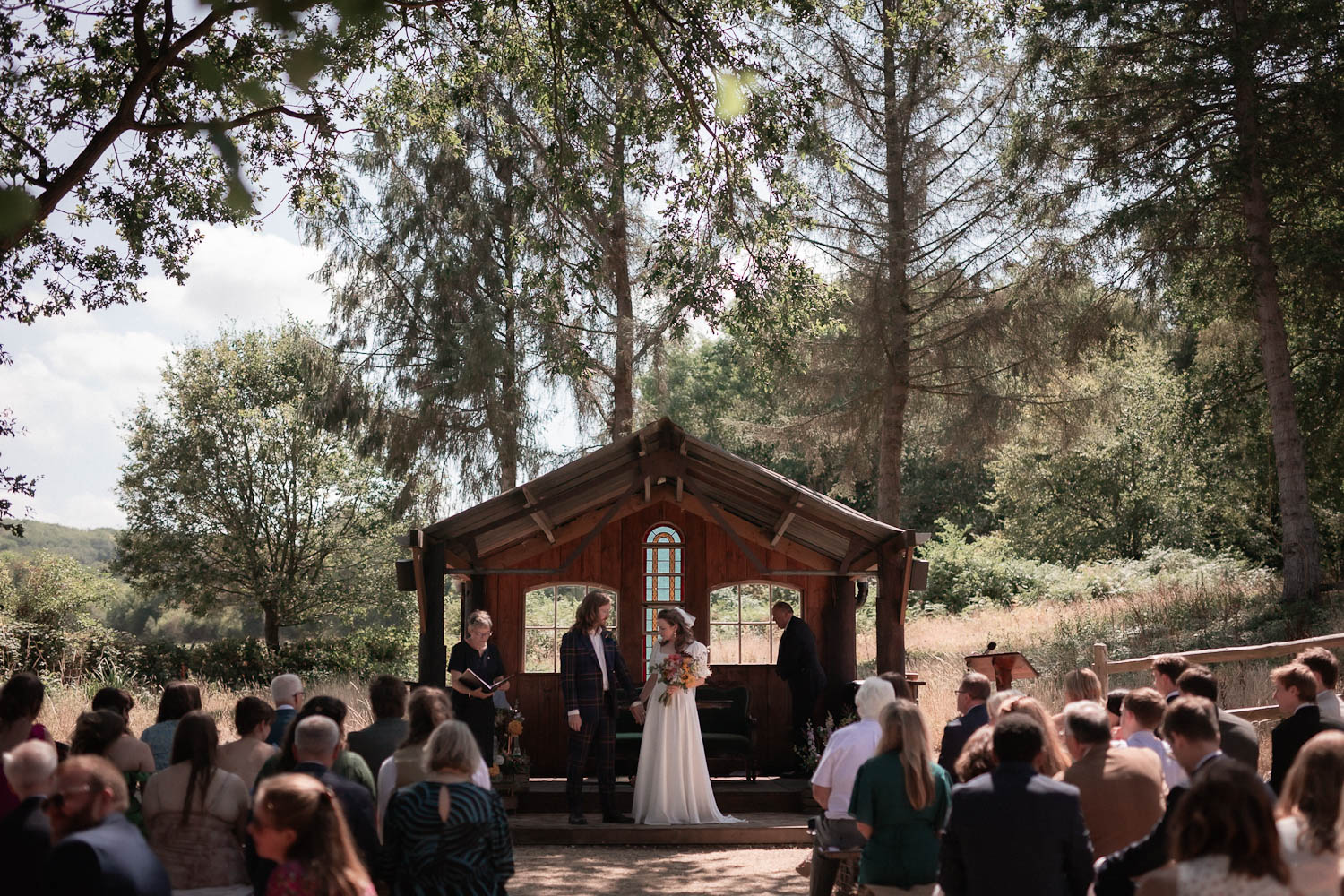 Outdoor wedding ceremony in front of a small wooden chapel, with guests seated and a couple standing at the altar among the trees.
