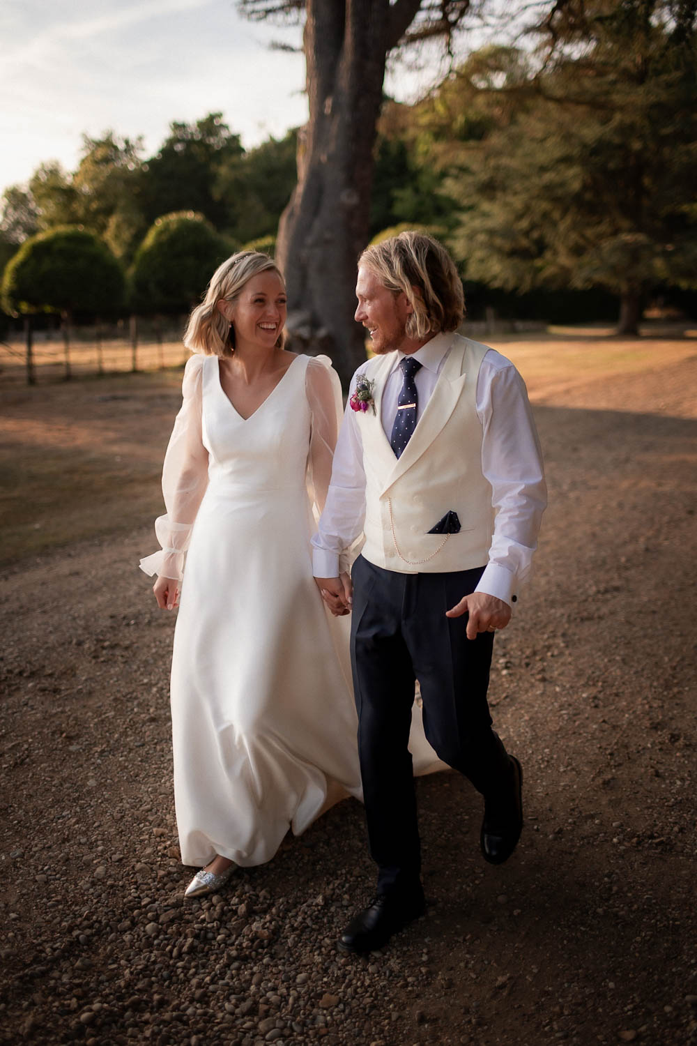 Bride and groom walking hand in hand along a woodland path in warm natural light.