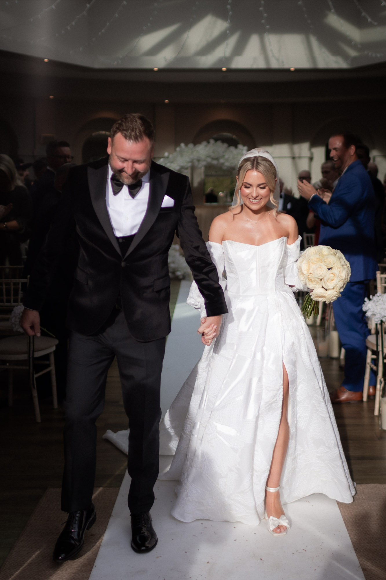 Bride and groom walking hand in hand down the aisle, smiling as guests clap during their wedding ceremony.