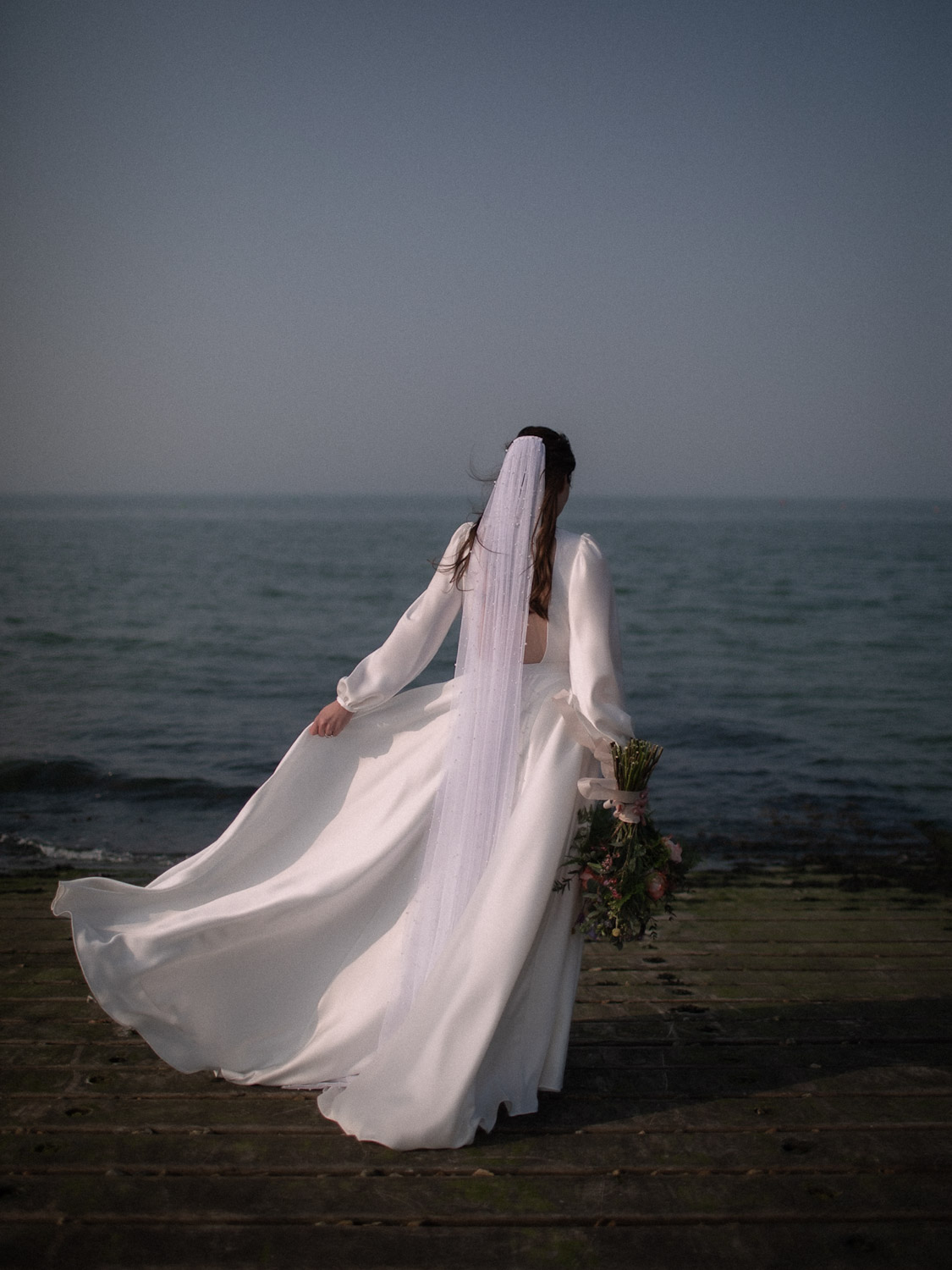 The bride stands alone on a wooden pier overlooking the sea, wearing a long white gown and veil and holding a pastel bouquet as the calm water stretches into the horizon.