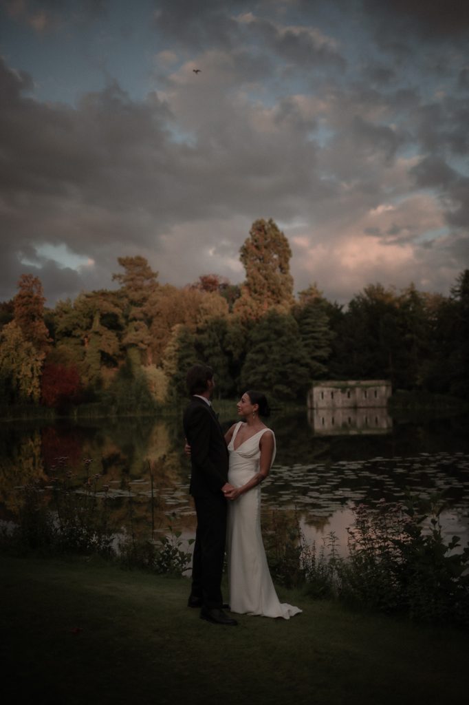 Bride and Groom at dusk stood in front of a gorgeous lake with a pink and blue sky. The trees are autumnul