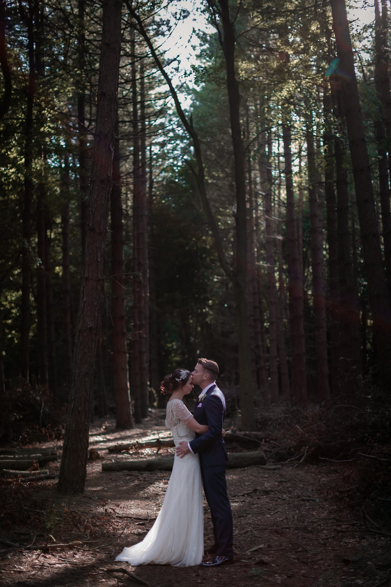 A couple embracing in a quiet woodland, the bride in a detailed white gown and the groom in a dark suit, surrounded by tall trees with soft sunlight filtering through the branches.