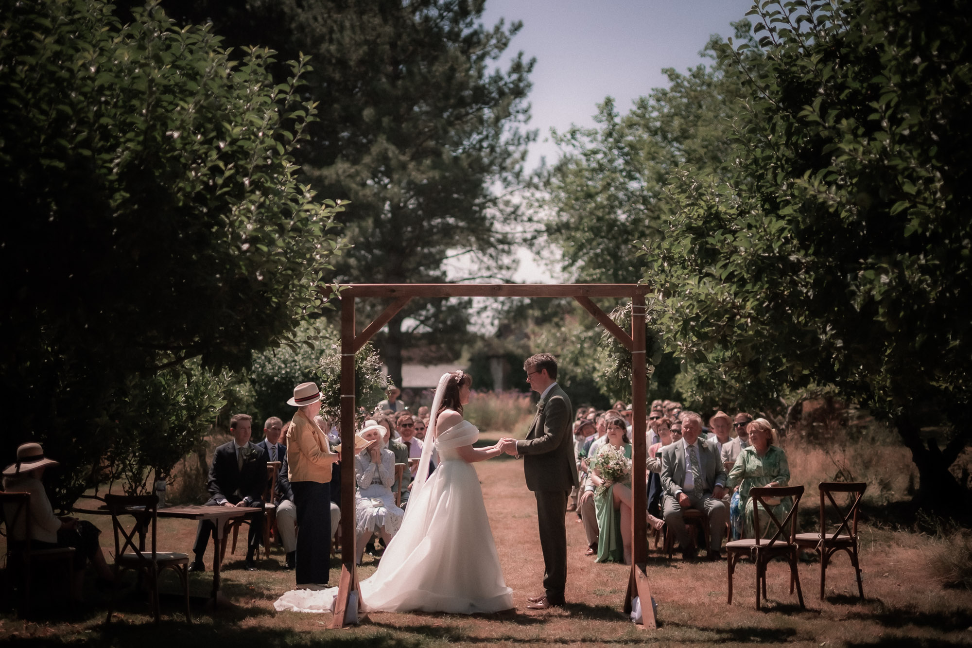 A bride and groom stand under a wooden arch decorated with greenery during an outdoor wedding ceremony, holding hands as guests watch from their seats.