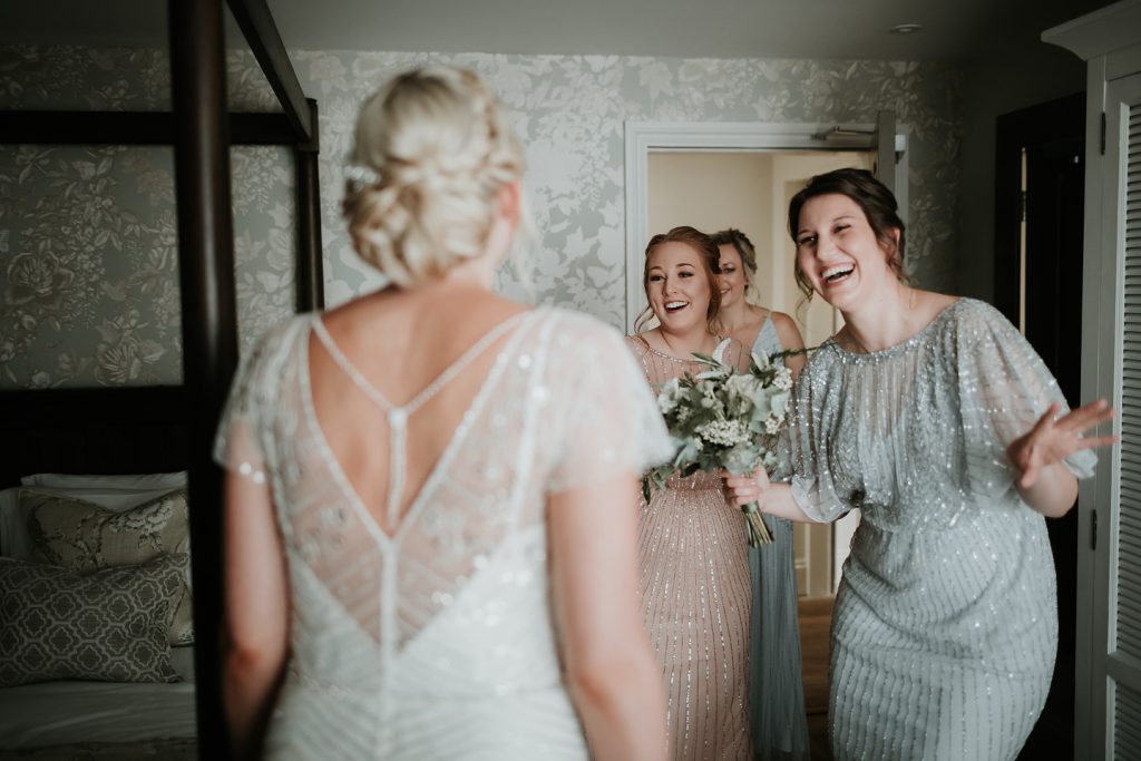 A bride in a beaded gown stands with her back to the camera while three bridesmaids in sequined dresses smile and react warmly in a bedroom. One bridesmaid holds a bouquet, and the group shares a joyful, candid moment before the wedding.
