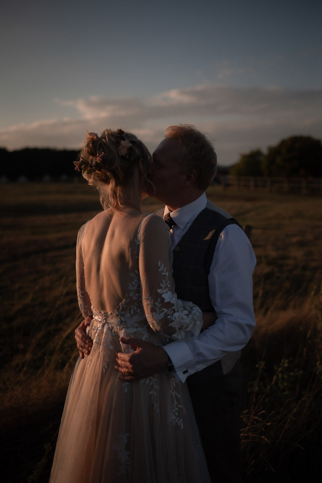 The couple embrace in a sunlit field at sunset, the bride in a lace-backed gown and floral hairpiece and the groom in a vest and tie, surrounded by tall grass and warm golden light.