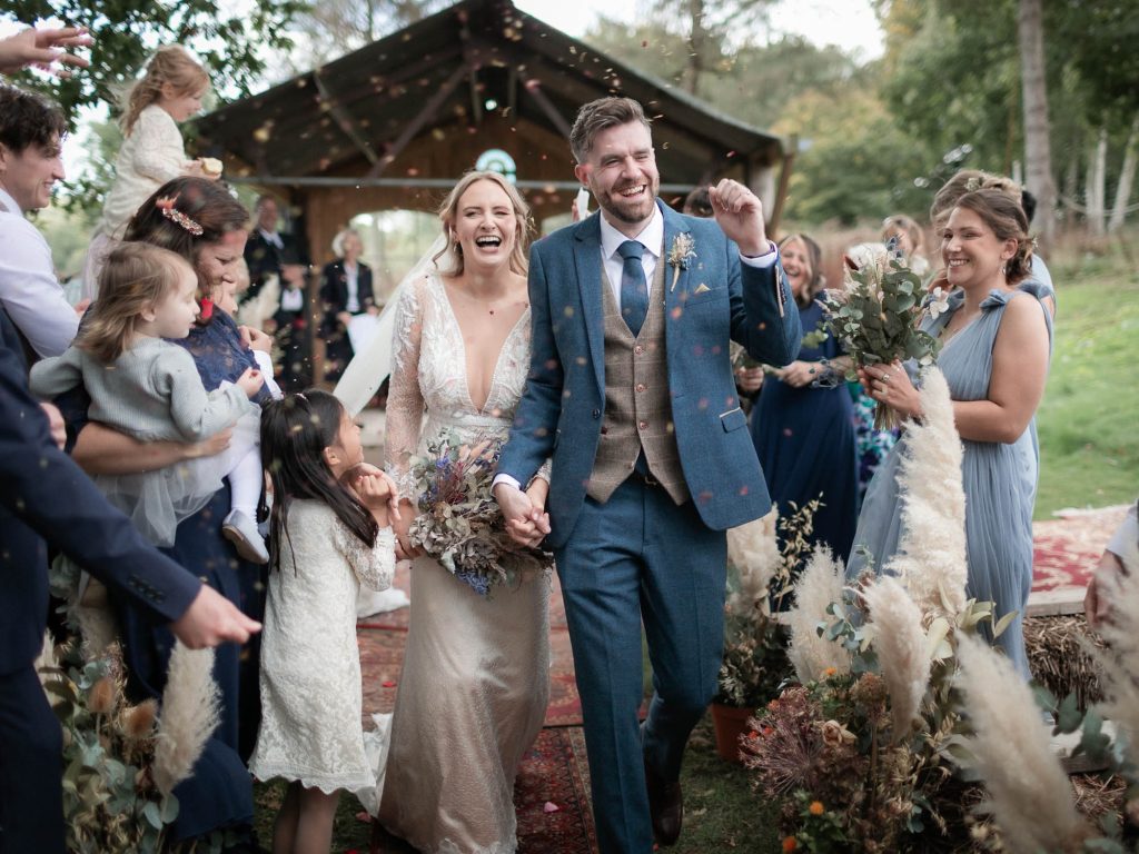 Bride and Groom walking through confetti tunnel, groom is cheering and holding his fist in the air and the bride laughing with a happy expression